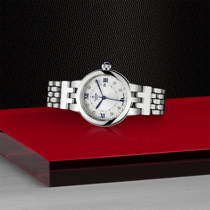 The TUDOR Clair de Rose, a silver luxury wristwatch with a white face, is displayed on a red and silver surface against a black background.