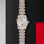 The TUDOR 1926 watch, featuring a silver and rose gold bracelet, white textured dial, rose gold bezel, and diamond hour markers, is displayed on a textured black and red background.
