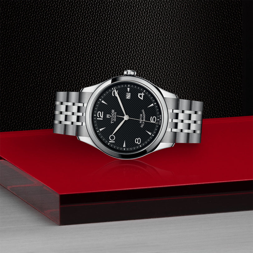 A TUDOR 1926 silver wristwatch with a black dial, silver hour markers, and metal bracelet is showcased on a glossy red and black surface against a textured black background—an elegant pick for mechanical watch enthusiasts.