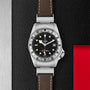 The TUDOR Black Bay P01 wristwatch, with signature snowflake hands, a brown leather strap, black dial, silver bezel, and date display, is displayed on a textured black and red surface.