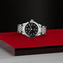 The TUDOR 1926, a timelessly elegant silver wristwatch with a black face and metal band, is showcased on a red and silver platform against a textured black background.