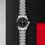 The TUDOR 1926 is a timelessly elegant stainless steel wristwatch featuring a black textured dial, silver hour markers, and a date display, displayed on a black textured and red geometric background.