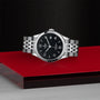 The TUDOR 1926, a classic stainless steel wristwatch with a black dial and silver bracelet, is showcased on a red and black platform against a textured dark background.