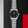 The TUDOR 1926 wristwatch, featuring a black dial, silver indices, and a stainless steel bracelet, is displayed on a textured black and metallic surface accented with red.