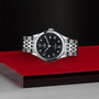 The TUDOR 1926 silver wristwatch with a black dial, silver markers, and metal bracelet rests on a red and silver surface against a textured black background—ideal for fans of classic mechanical timepieces.