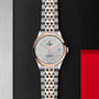 The TUDOR 1926 wristwatch features a silver and rose gold bracelet, silver dial, rose gold markers and hands, plus a date display—presented on a textured black and red background for classic watch enthusiasts.