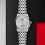 A TUDOR 1926 wristwatch in silver with a white textured dial, orange numerals and hands, and a metal link bracelet is displayed against a black and red background—showcasing TUDORs signature mechanical watch design.