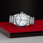 A TUDOR 1926 stainless steel wristwatch with a white dial, blue hour markers, and a metal bracelet is displayed on a glossy red and black surface.