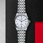 A TUDOR 1926 silver wristwatch featuring a white dial with blue numerals, date display, and silver metal bracelet, shown on a textured black background with red and silver accents.