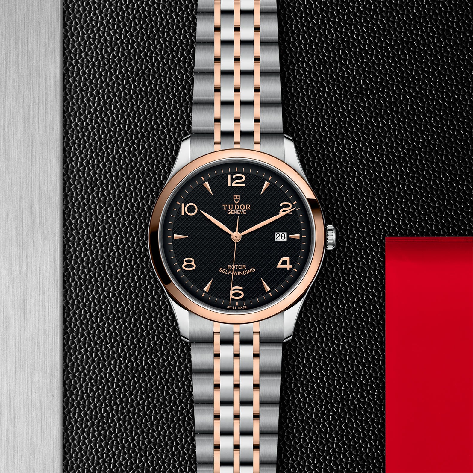 The TUDOR 1926 wristwatch, featuring stainless steel and rose gold with a black dial, gold hour markers, and date display, is displayed on a textured black and red background—an elegant mechanical timepiece for any collection.