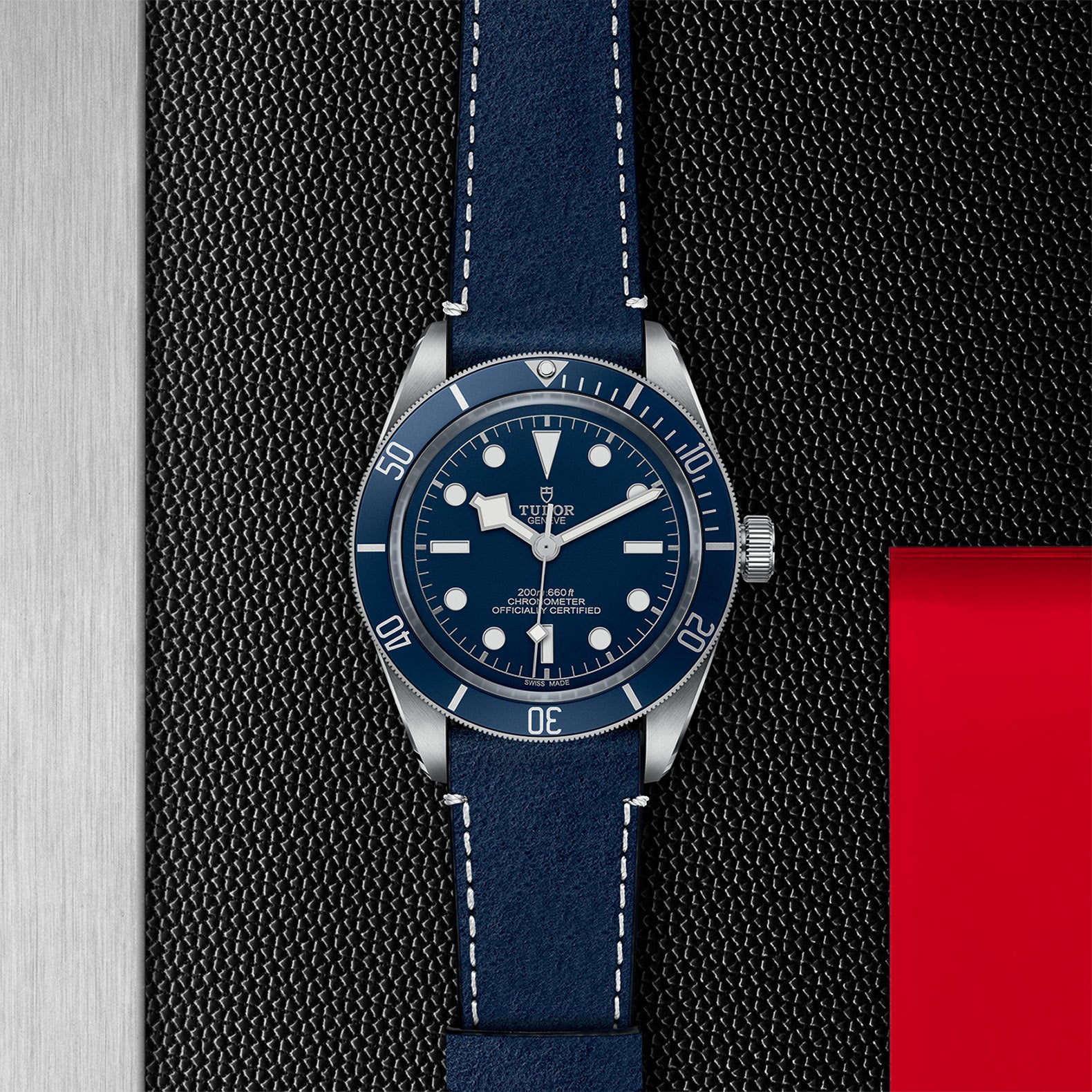 A TUDOR Black Bay 58 wristwatch with a blue dial and leather strap rests on a textured black surface, featuring metal and red accents.