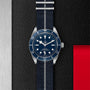 The TUDOR Black Bay 58, a blue diving watch on a striped fabric strap, is shown on a textured black surface with silver metal and red accents, highlighting its self-winding mechanical movement.