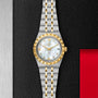 The TUDOR Royal two-tone wristwatch, featuring a stainless steel and gold band, white dial, gold Roman numerals, and diamond hour markers, is showcased against a textured black and red backdrop.
