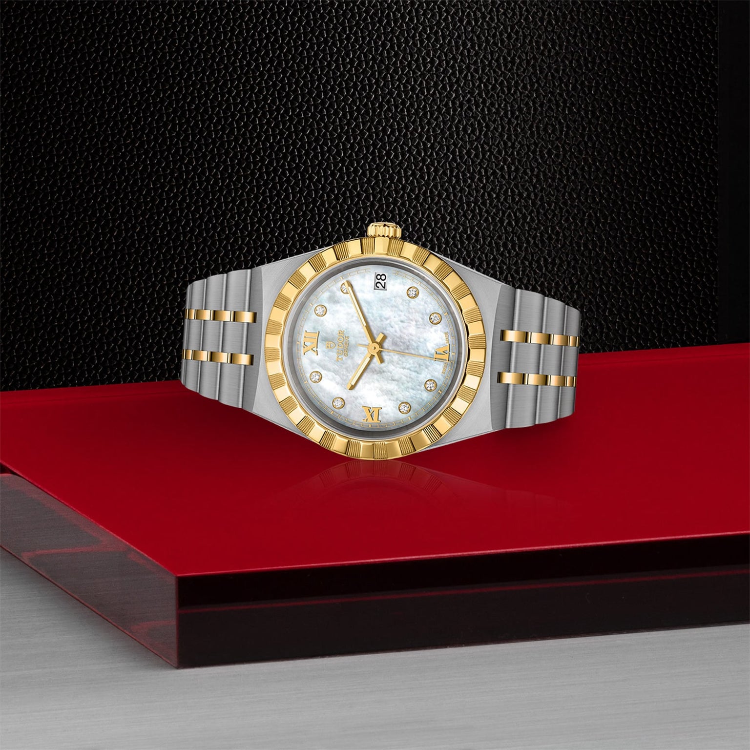 A TUDOR Royal wristwatch with a gold bezel and hour markers, white dial, and two-tone stainless steel and gold bracelet is displayed on a red and silver surface against a textured black background.