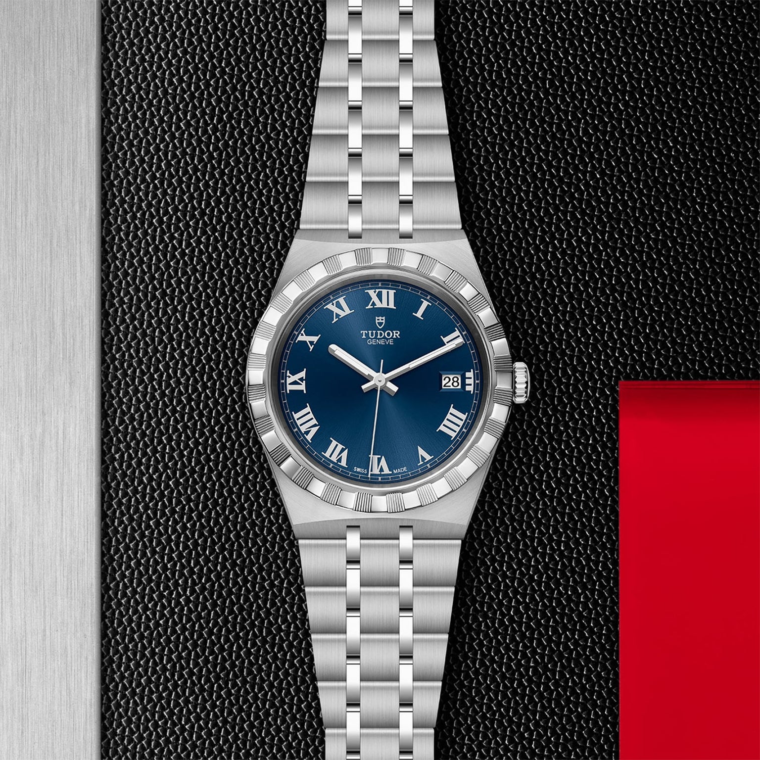 The TUDOR Royal is a stainless steel wristwatch with a blue dial, Roman numerals, and date display, showcased on a textured black surface with red and silver accents—highlighting its automatic sport-chic style.