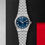 The TUDOR Royal is a stainless steel wristwatch with a blue dial, Roman numerals, and date display, showcased on a textured black surface with red and silver accents—highlighting its automatic sport-chic style.