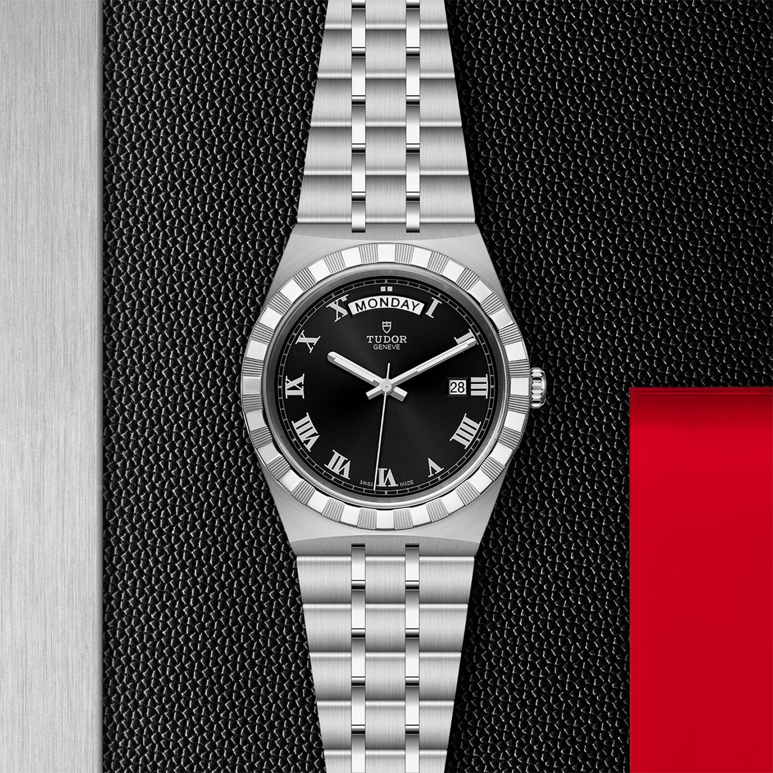 The TUDOR Royal, a stainless steel wristwatch with black dial, Roman numerals, and day-date display, exemplifies automatic sport-chic style against a textured black and red backdrop.