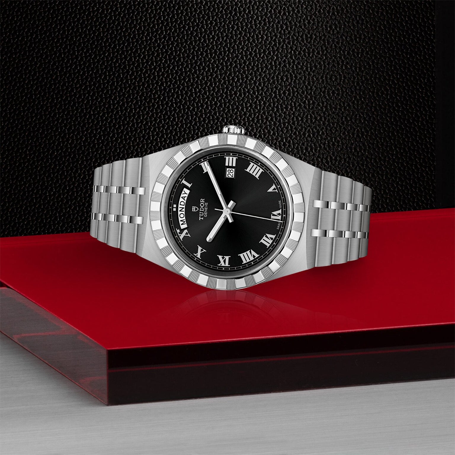 The TUDOR Royal, a silver automatic sport-chic watch with a black dial, Roman numeral markers, and a metal bracelet, is displayed on a red and black surface.