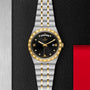 The TUDOR Royal watch features a black dial with gold accents and a two-tone silver and gold integrated bracelet, elegantly displayed on a textured black and silver background with red—highlighting its automatic sport-chic design.