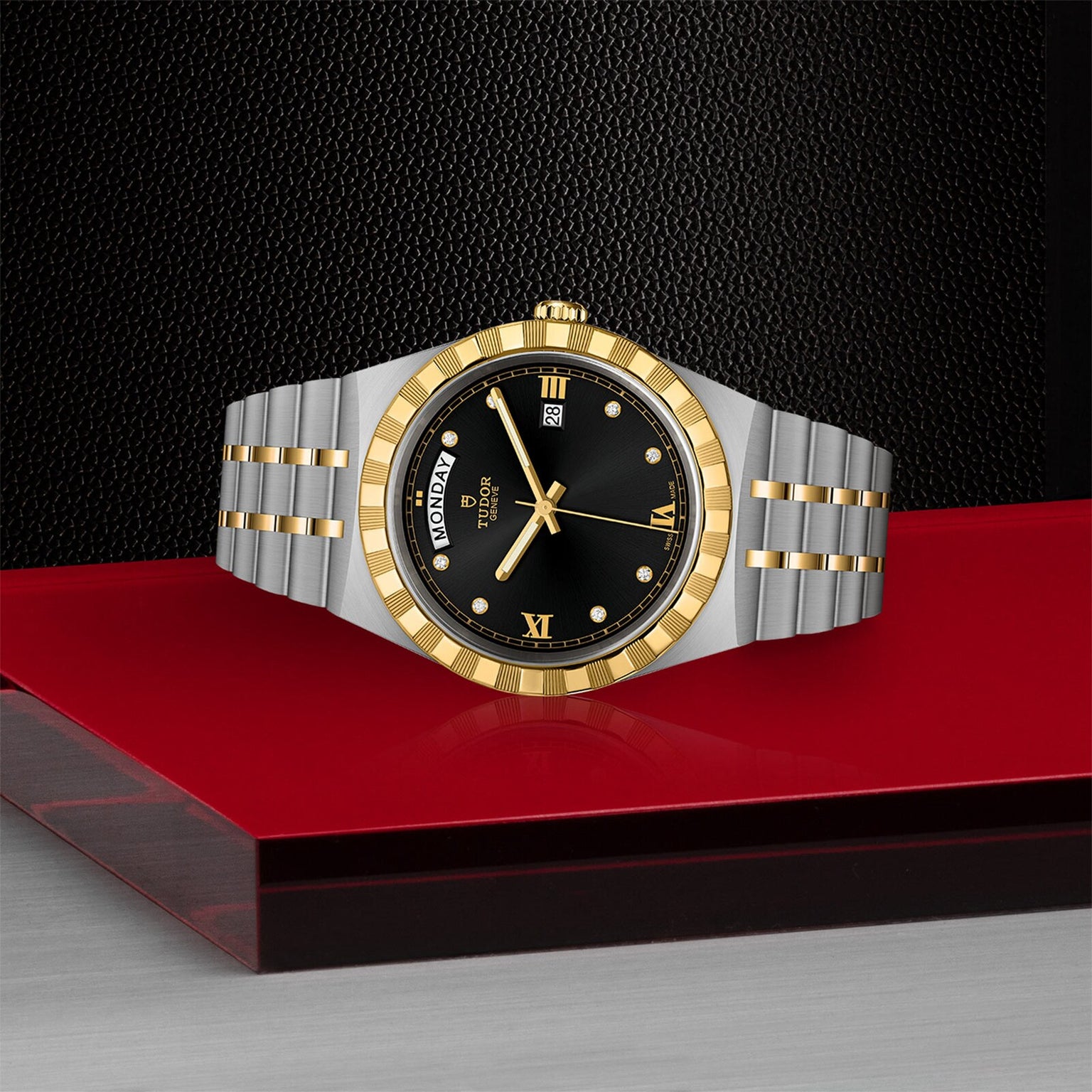 A TUDOR Royal wristwatch with a silver and gold finish, black dial, and integrated bracelet is showcased on a red and gray platform against a textured black background.