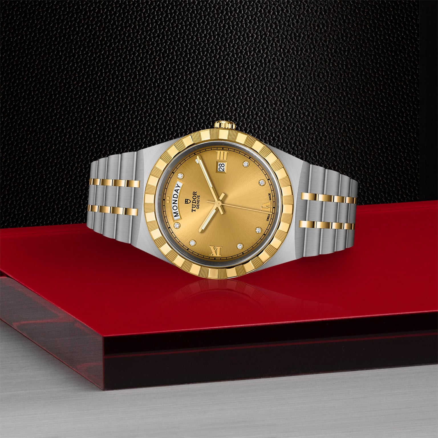 A TUDOR Royal wristwatch in stainless steel and gold, with a gold dial, Roman numerals, and day-date display, rests elegantly on a glossy red and black surface.
