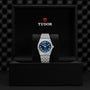 A TUDOR Royal watch with a blue dial and silver metal bracelet stands upright in a black Tudor-branded presentation box.