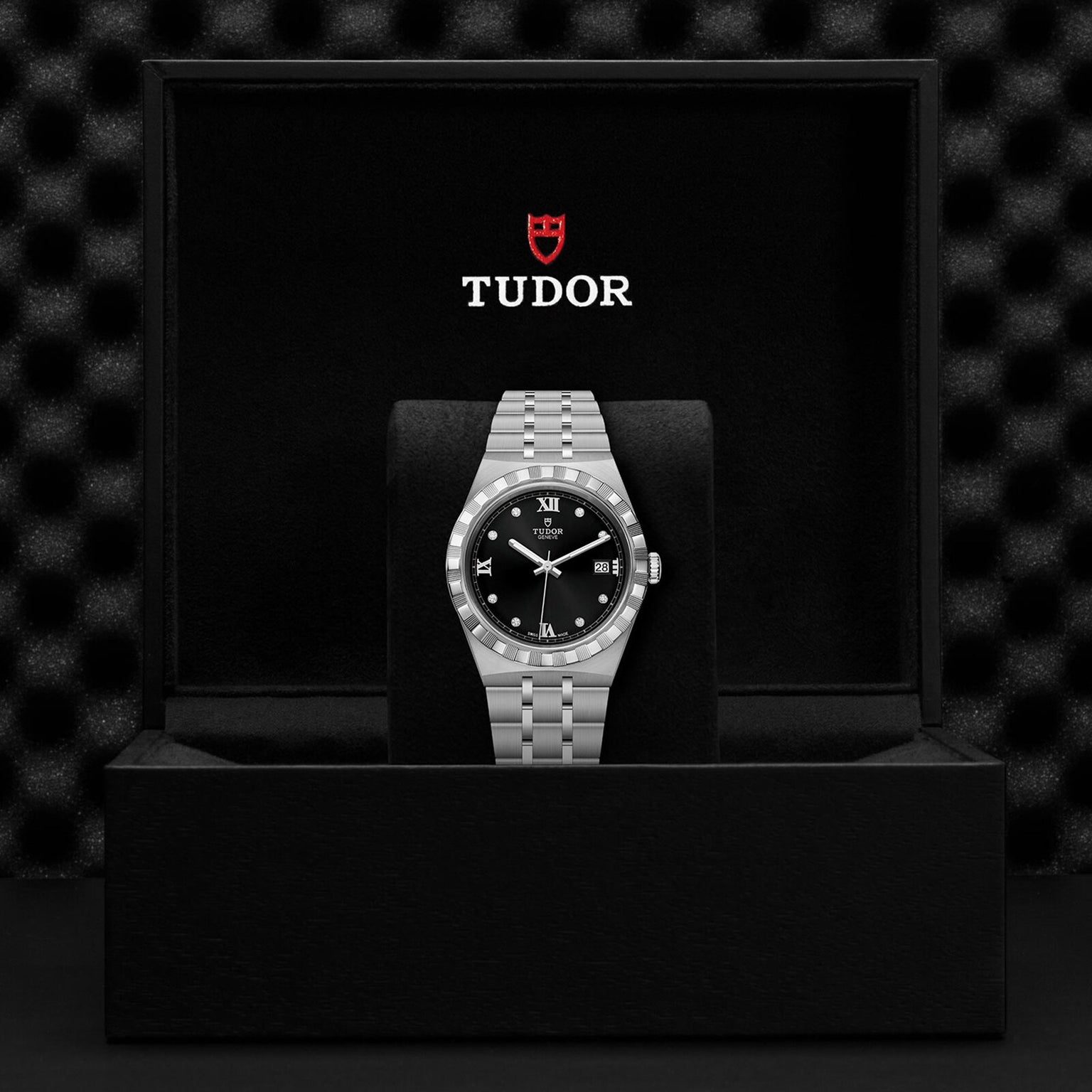 A silver TUDOR Royal wristwatch with a black dial is shown standing in an open black TUDOR box, set against a textured black background that highlights the automatic sport-chic appeal of the Royal collection.