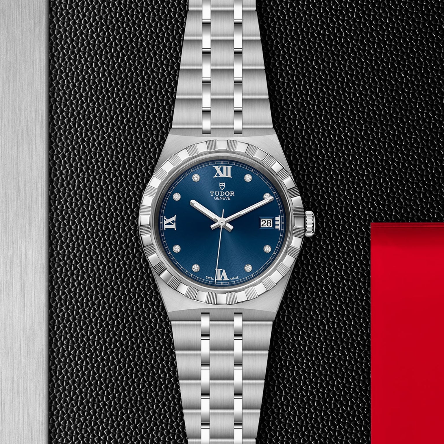 The TUDOR Royal wristwatch features a blue dial with Roman numerals, date display, and an integrated bracelet, shown on a black and metal surface with a red accent—an ideal automatic sport-chic choice for watch enthusiasts.