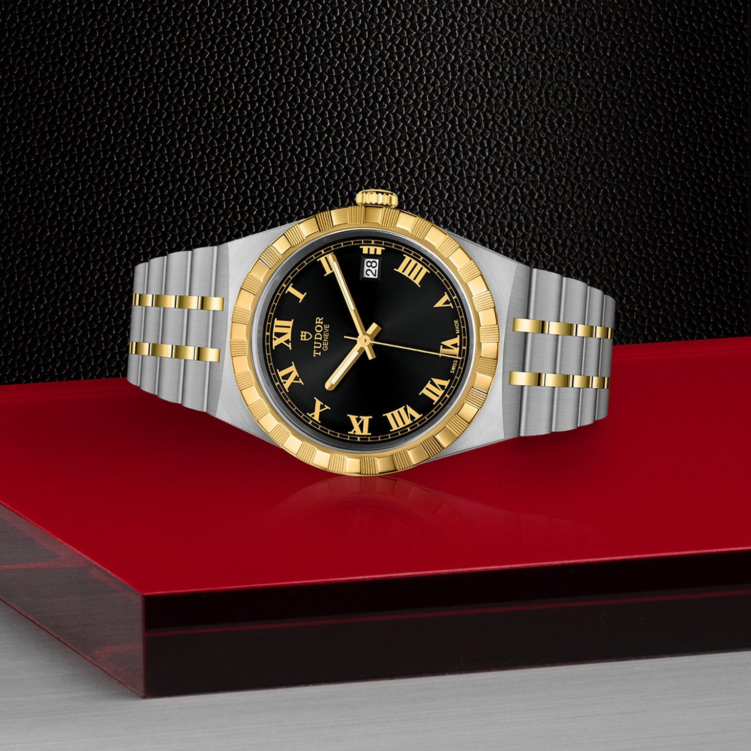 A TUDOR Royal wristwatch with a gold and silver finish, black dial, Roman numerals, date display, and integrated bracelet is showcased on a red and glass surface against a textured black backdrop—exemplifying sport-chic style.