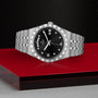 The TUDOR Royal stainless steel wristwatch features a black dial showing day, date, and time. Displayed on a red and transparent surface with a textured black background, its an elegant choice in the sport-chic watch category.