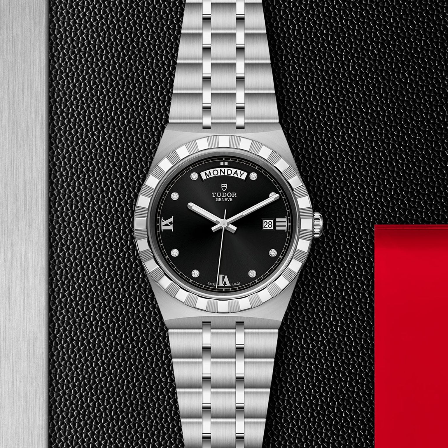 The TUDOR Royal wristwatch, featuring a black dial, day and date display, and silver bracelet, rests on a textured black and metal surface with a red accent—showcasing the sport-chic elegance of TUDORs automatic watches.