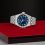 The TUDOR Royal, a silver wristwatch with a blue face and Roman numerals, is displayed on a red and black textured surface.