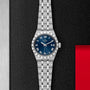 A TUDOR Royal wristwatch with a silver case, blue dial, and Roman numerals is displayed on a textured black surface.