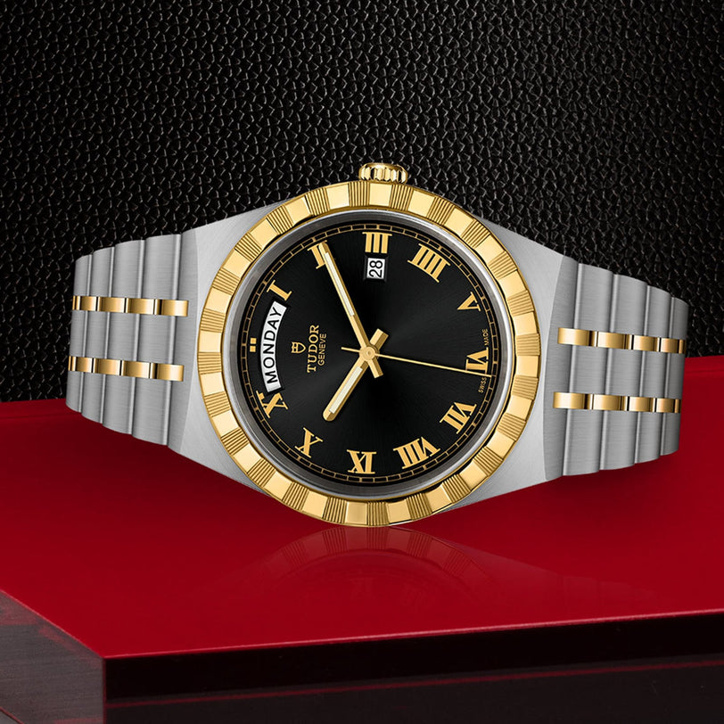 A TUDOR Royal wristwatch with a black dial, gold Roman numerals, and day-date display on an integrated bracelet is shown on a textured red and black surface.