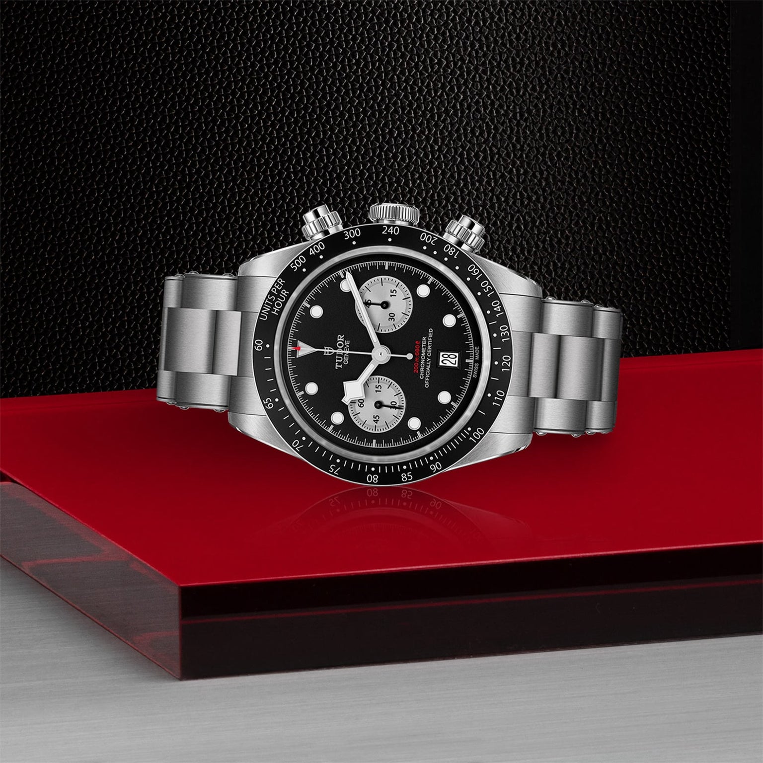 The TUDOR Black Bay Chrono, a stainless steel wristwatch with a tachymetric scale, black bezel, and black dial, is showcased on a glossy red and silver platform against a dark textured background.
