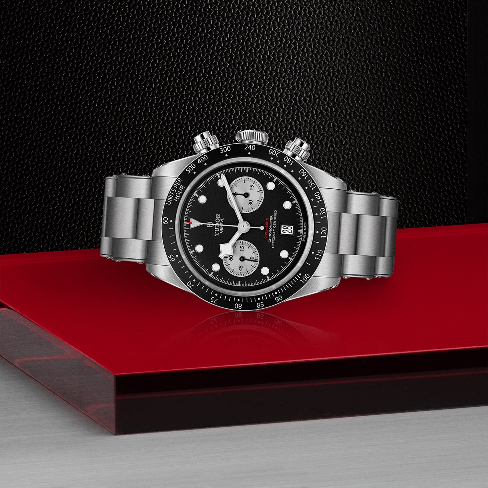 The TUDOR Black Bay Chrono, a stainless steel wristwatch with a tachymetric scale, black bezel, and black dial, is showcased on a glossy red and silver platform against a dark textured background.