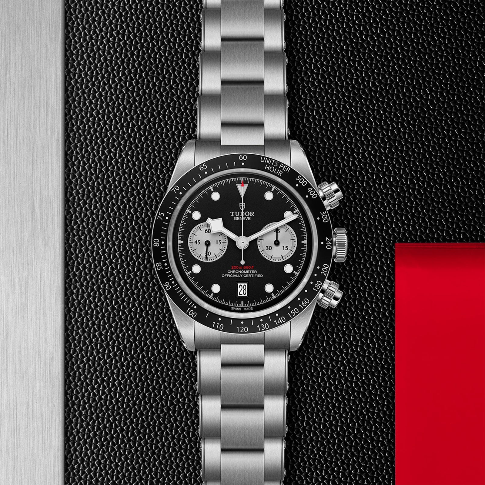 A TUDOR Black Bay Chrono stainless steel watch, featuring a black dial and tachymetric bezel, rests on a dark textured surface beside a red rectangular accent.