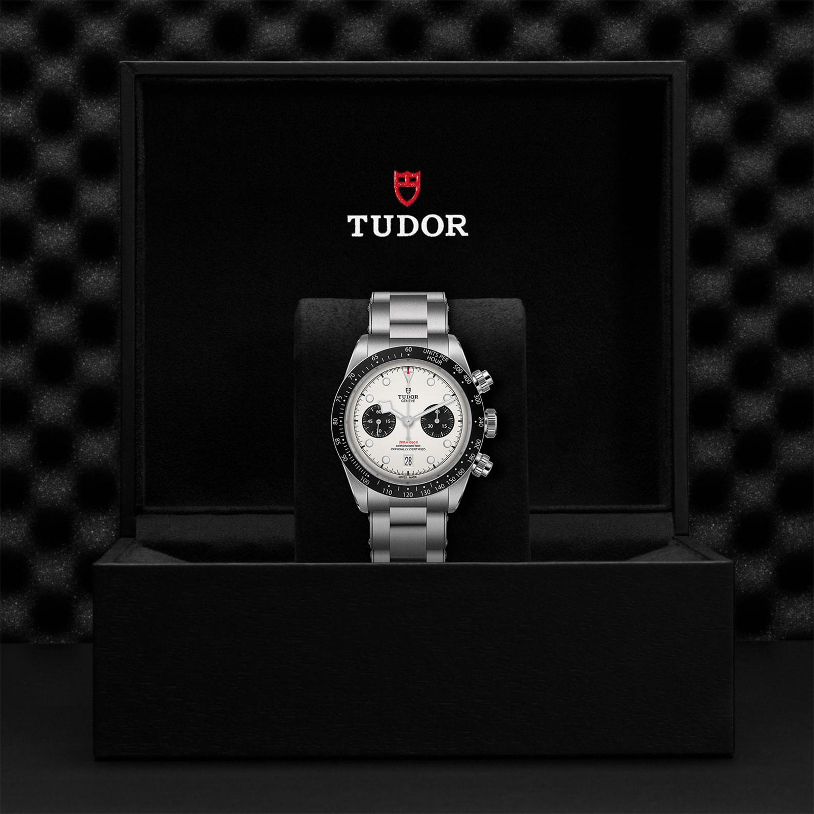 The TUDOR Black Bay Chrono wristwatch with a silver band is displayed in a black TUDOR box featuring the brand’s logo.
