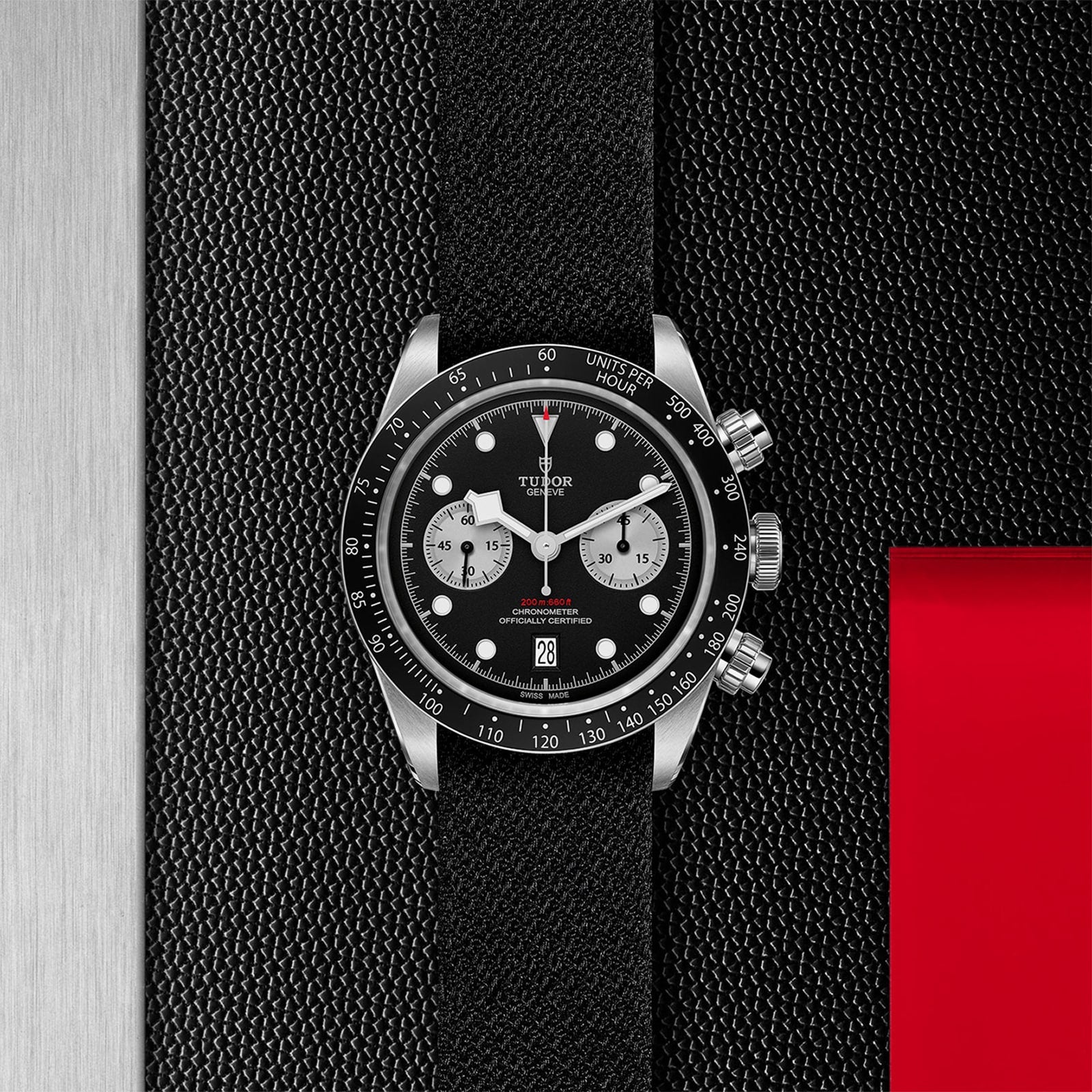 The TUDOR Black Bay Chrono features a black bezel, black textured strap, and two silver subdials, set against a black textured background with bold red and silver accents—a striking chronograph from TUDOR.