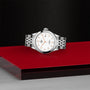 A TUDOR 1926 silver wristwatch with a white dial and metal bracelet is showcased on a red and black platform against a textured black background.