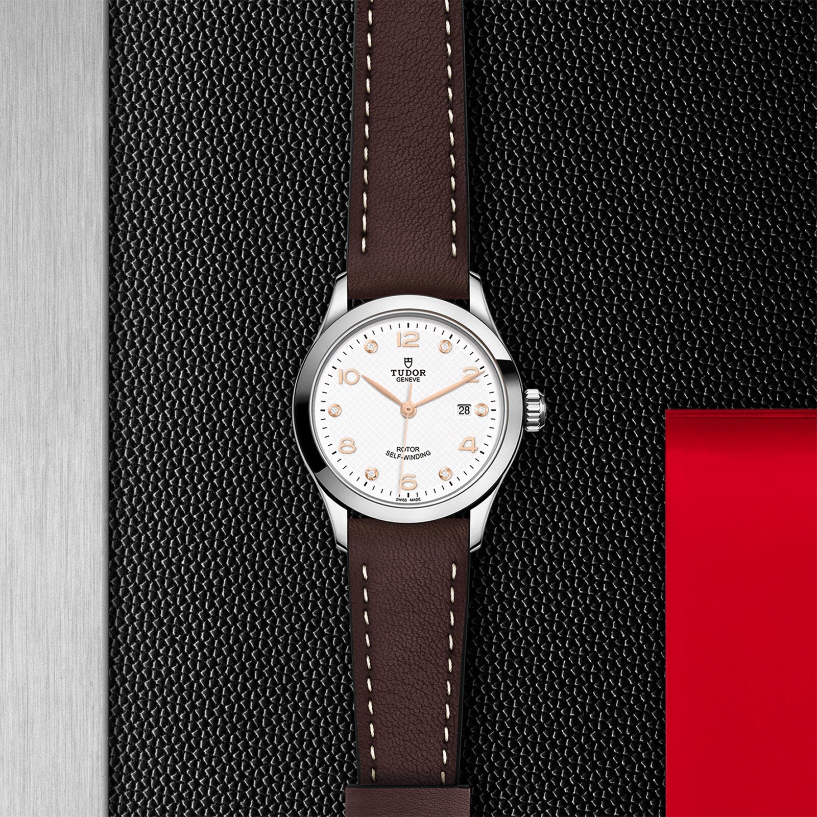 The TUDOR 1926 wristwatch features a brown leather strap and a white dial, set against a textured black and silver background.
