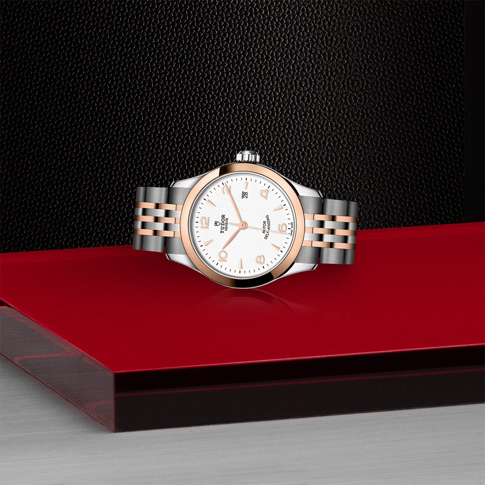 The TUDOR 1926, an elegant two-tone wristwatch with a silver and rose gold bracelet, is displayed on a glossy red-and-black platform set against a textured dark background.