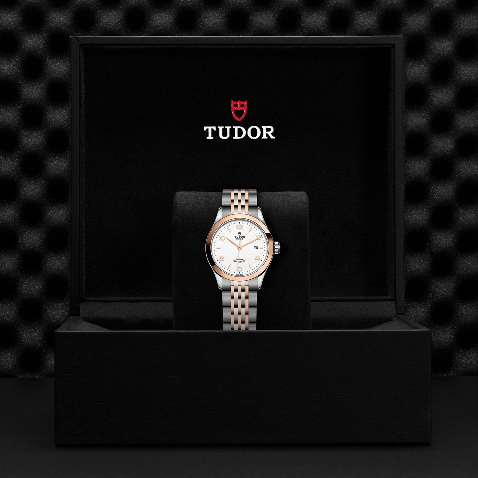 The TUDOR 1926 wristwatch, with a silver and gold bracelet, is elegantly showcased upright in a black box featuring the TUDOR logo on the inside lid.
