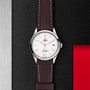A TUDOR 1926 wristwatch featuring a white dial, brown leather strap, silver bezel, and date display rests on a textured black surface with red and metallic details—perfect for fans of classic elegance.