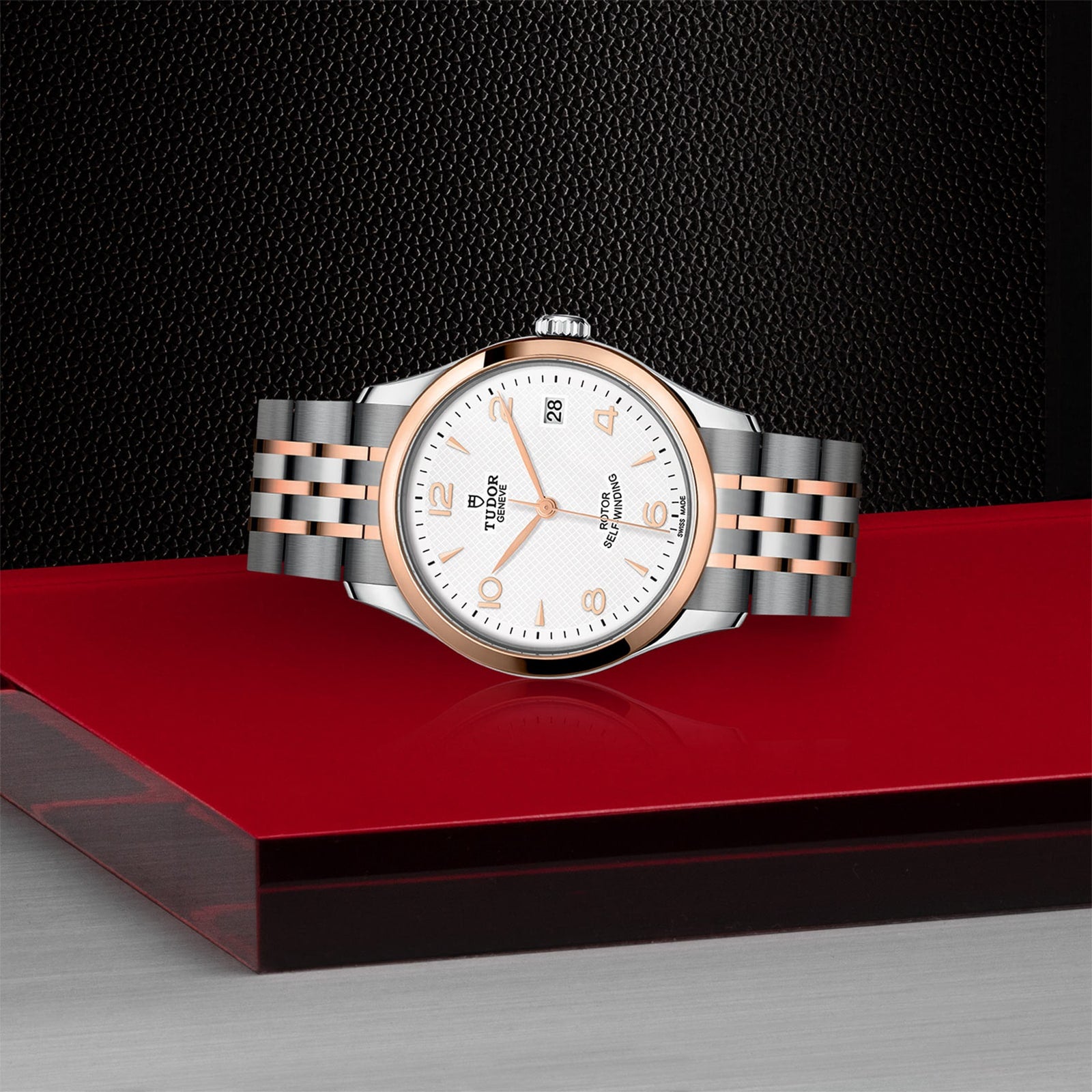 The TUDOR 1926 wristwatch features a silver and rose gold bracelet and white dial, showcased against a red and black surface with a textured black background.