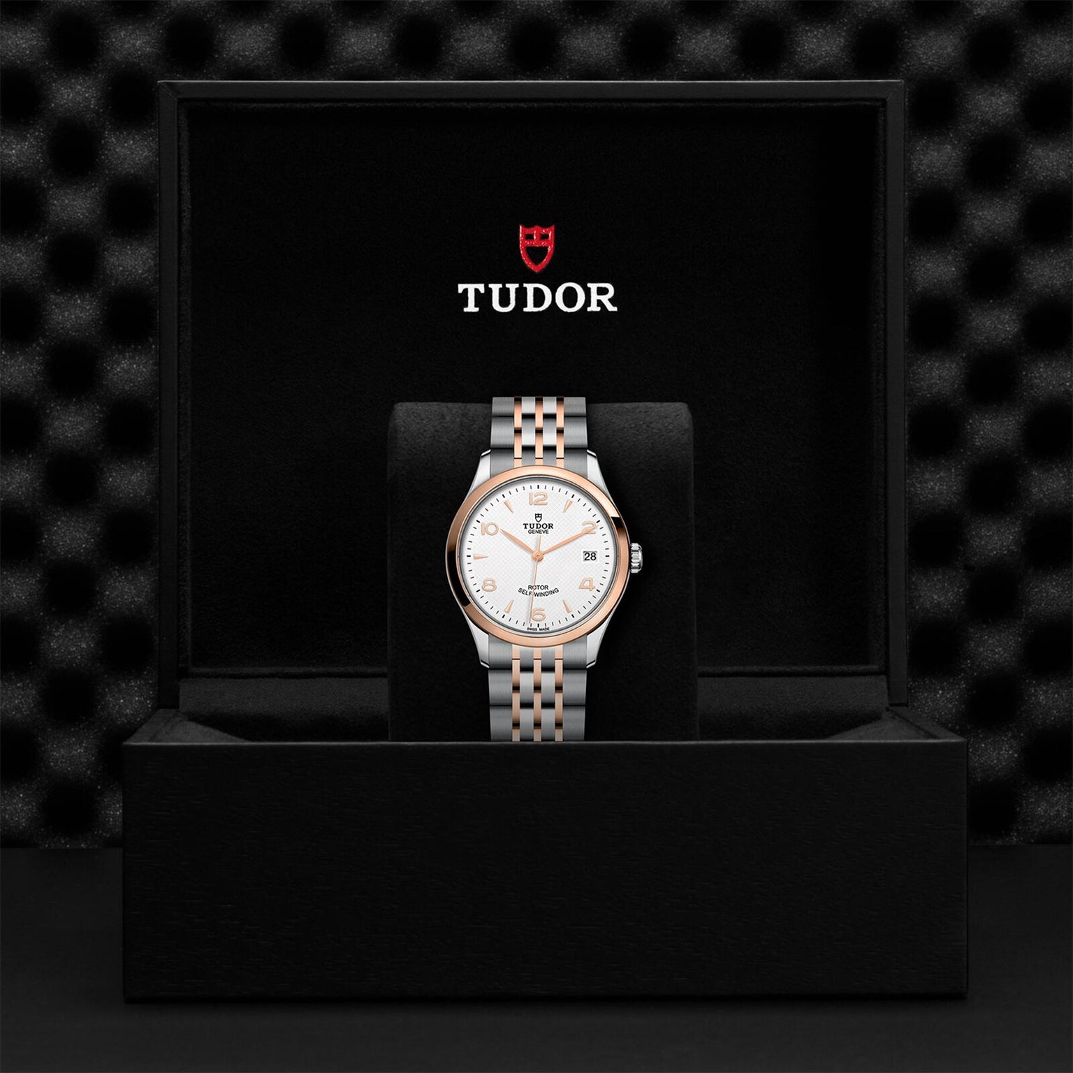 A TUDOR 1926 wristwatch with a white dial and two-tone metal band is displayed upright in an open black TUDOR box with the brand logo on the inside lid, emphasizing the heritage of mechanical watchmaking.