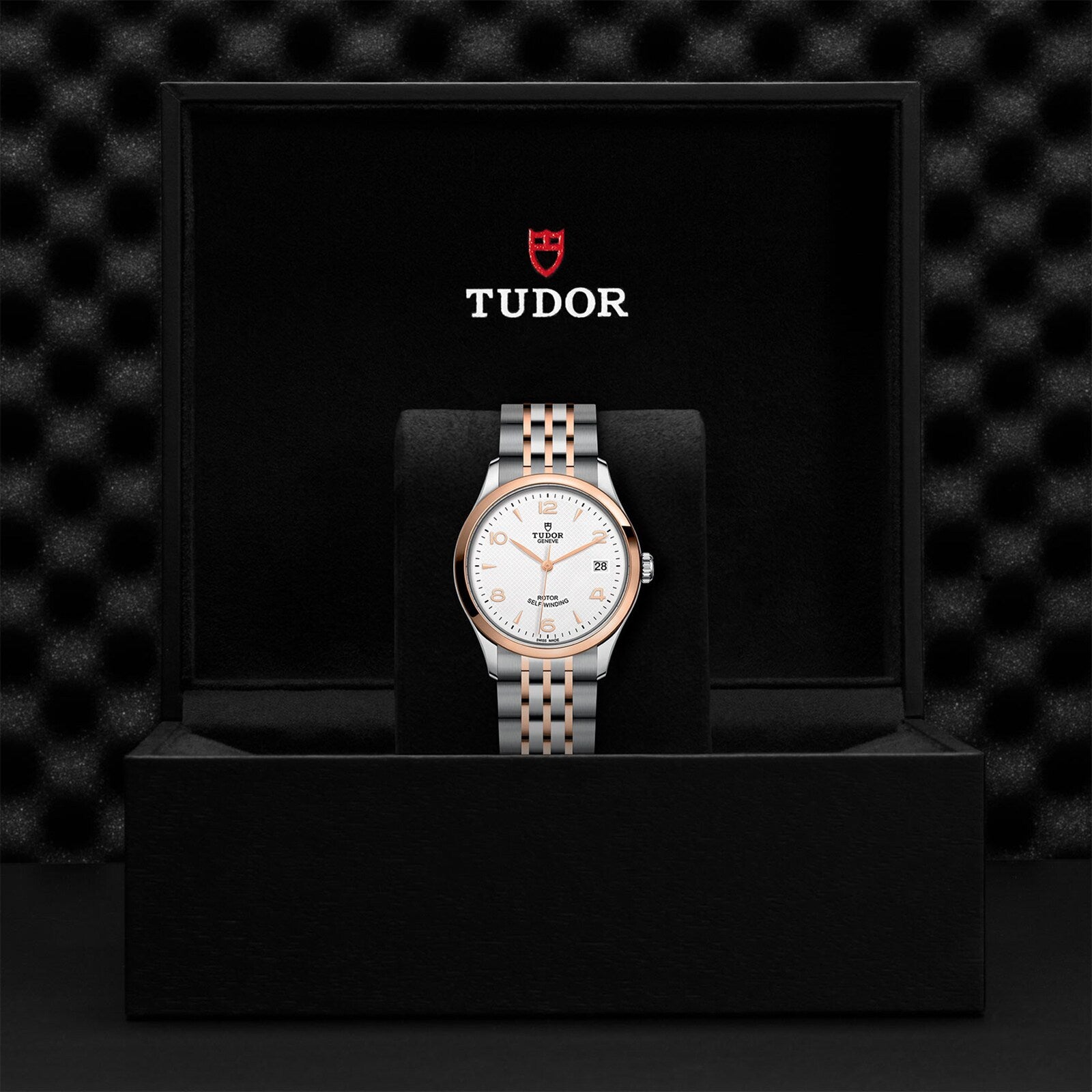 A TUDOR 1926 wristwatch with a white dial and two-tone metal band is displayed upright in an open black TUDOR box with the brand logo on the inside lid, emphasizing the heritage of mechanical watchmaking.