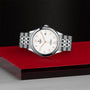 The TUDOR 1926, a silver wristwatch with a white dial, gold hour markers, and date display—symbolizing classic elegance—is displayed on a striking red and black geometric platform.