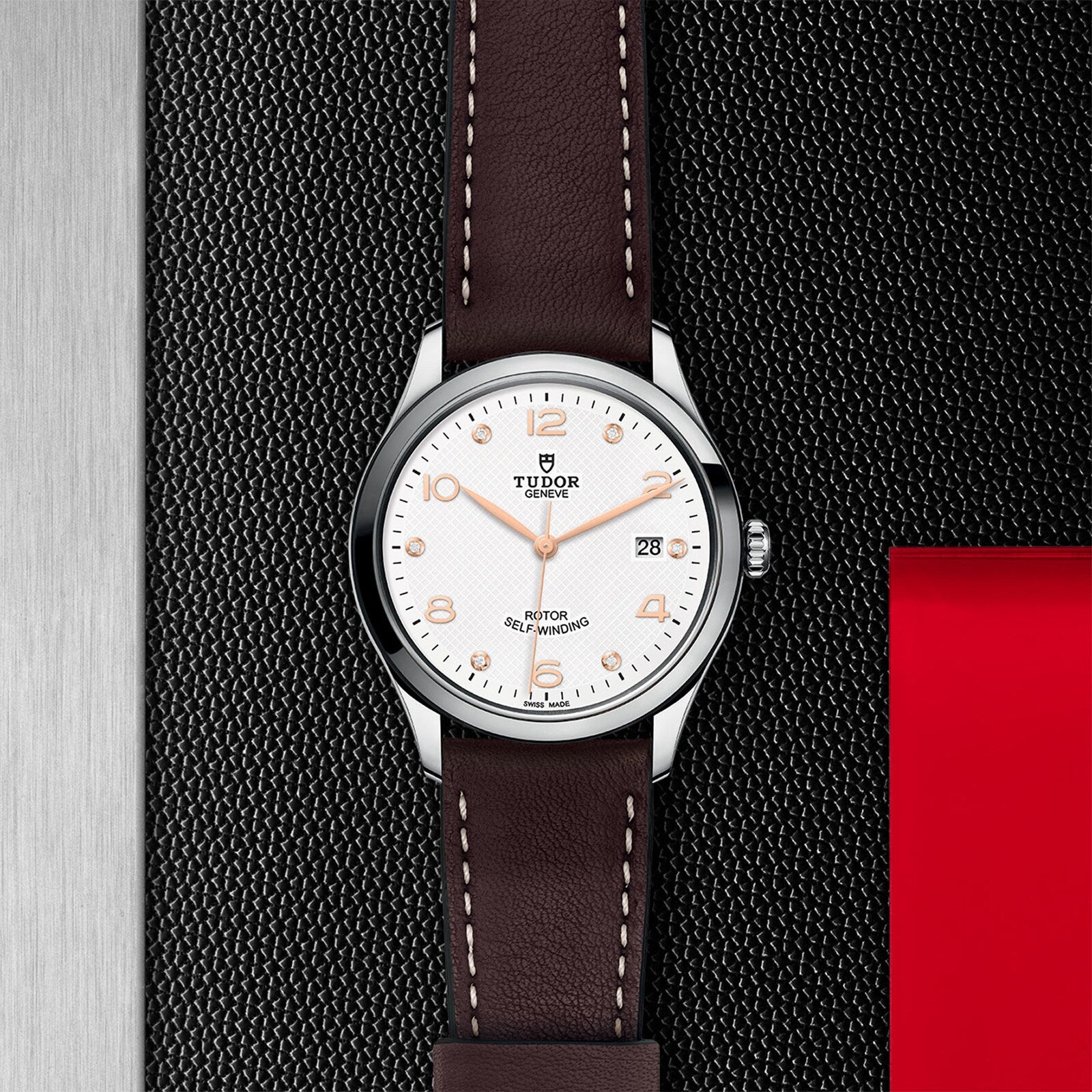A TUDOR 1926 wristwatch with a brown leather strap and white dial displayed on a textured black and red background.