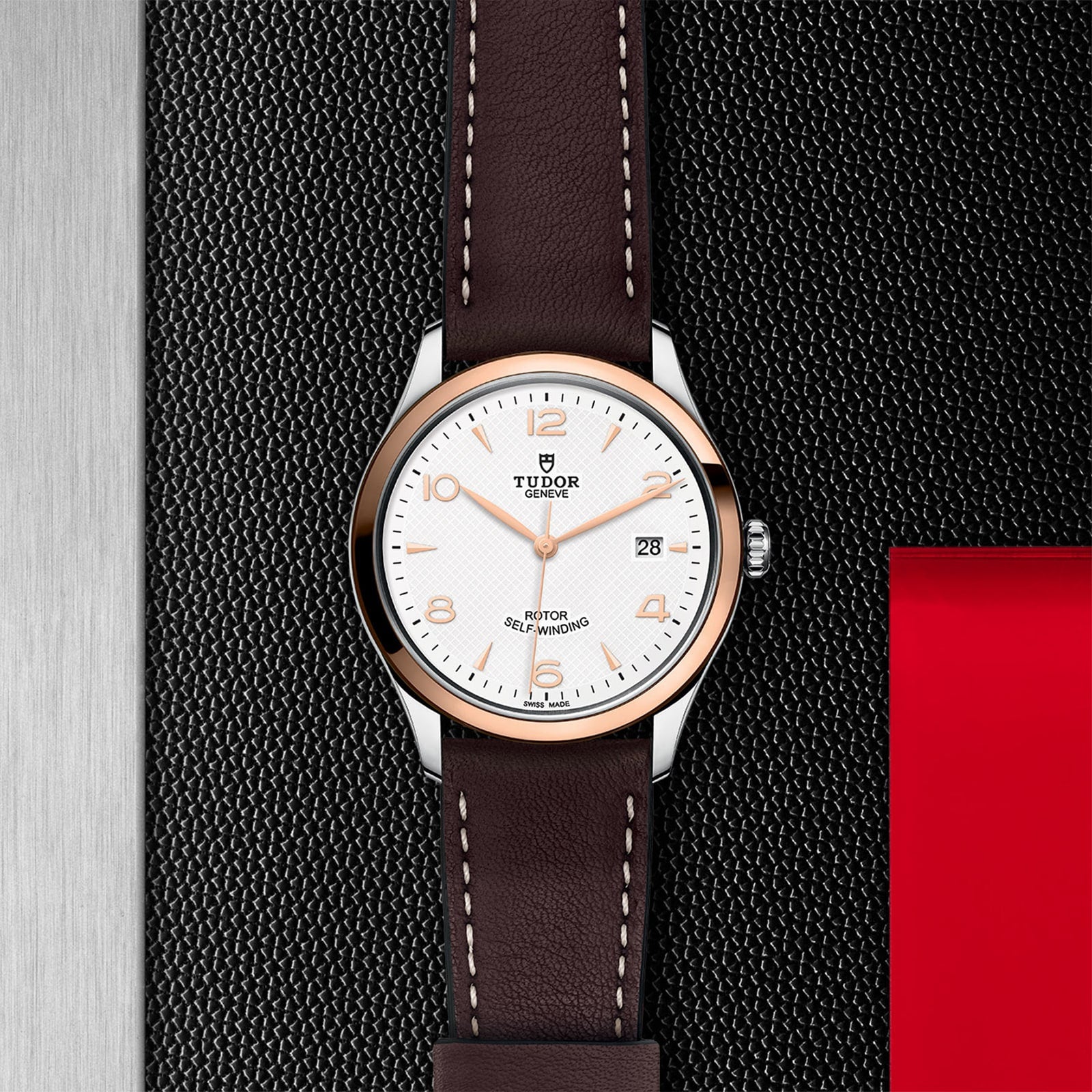 A TUDOR 1926 watch with a white dial, gold accents, brown leather strap, and date display sits on a textured black surface beside metal and red details—a classic choice for mechanical watch enthusiasts.